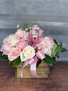 Floral arrangement with pink and white flowers, a bird figurine, and a pink ribbon in a wooden box on a wooden surface.