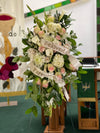 Traditional funeral standing spray with white and pink flowers, greenery, and ribbons reading “Beloved Parents”