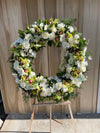 Standing funeral wreath with white roses, green cymbidium orchids, and burgundy accents on a wooden easel